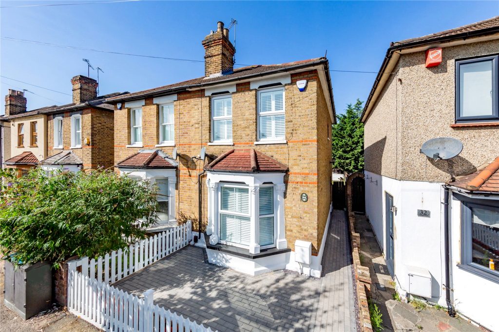 A London stock brick semi-detached house on Cotleigh Road with red brick banding, a tiled canopy over the bay window and plantation shutters, behind a white picket fence